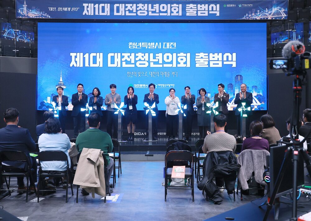 무대 위에 남녀 여러 사람이 일렬로 서서 박수를 치고 있고, 무대 앞 의자에 사람들이 앉아 있는 행사 장면