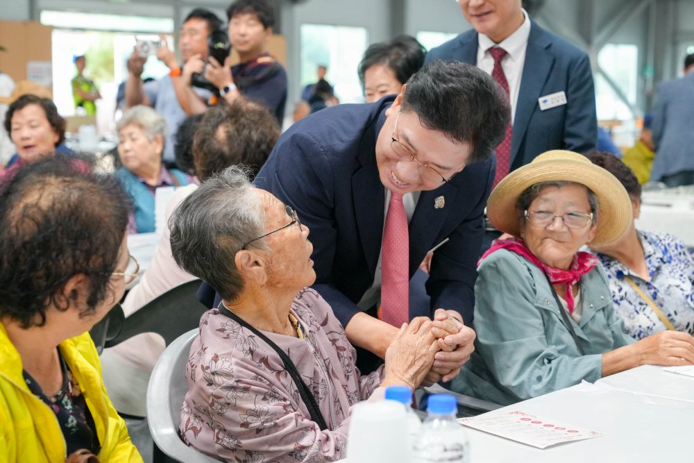 실내에서 여러 노인이 앉아 있고 남성이 한 노인의 손을 잡고 이야기를 나누는 모습