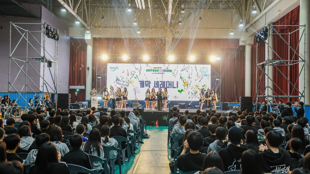 많은 관객이 좌석에 앉아 있고 무대 위에서 여러 사람이 서서 행사를 진행하는 실내 공간 전경