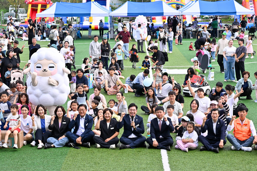 잔디 위에 앉은 어린이와 어른들이 밝게 웃으며 모여 있고 뒤쪽에 푹신한 양 캐릭터와 텐트가 보이는 장면