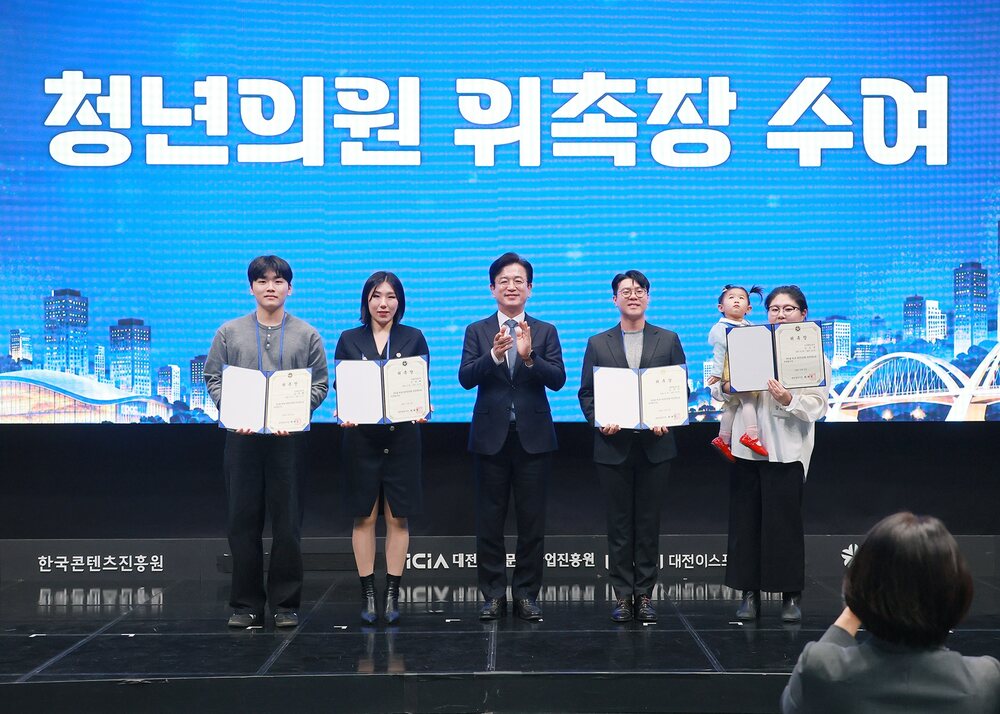 무대 위에 다섯 사람이 나란히 서서 상장과 위촉장을 들고 있으며 뒤쪽 스크린에 '청년의원 위촉장 수여'라고 적힌 행사 장면