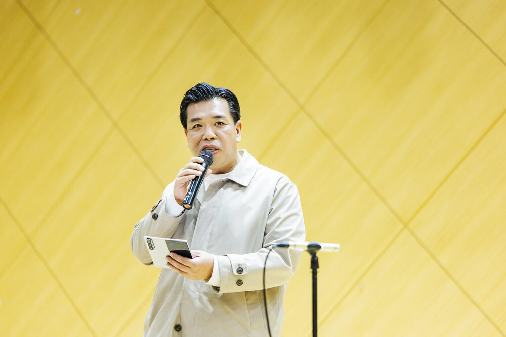 마이크를 잡고 휴대폰을 든 남성이 체크무늬 배경 앞에서 연설하는 모습