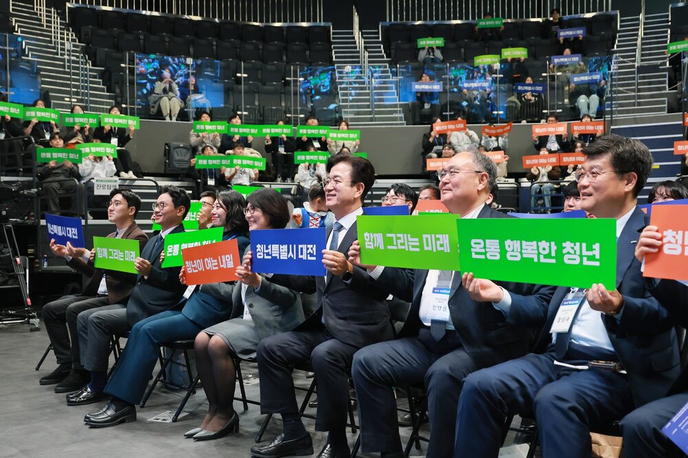 행사장 좌석에 앉아 다양한 문구가 적힌 손팻말을 들고 있는 여러 남녀 인물 모습