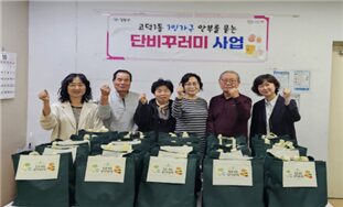 실내에서 여섯 명이 단비꾸러미 물품 앞에 서서 손가락 하트를 만드는 모습